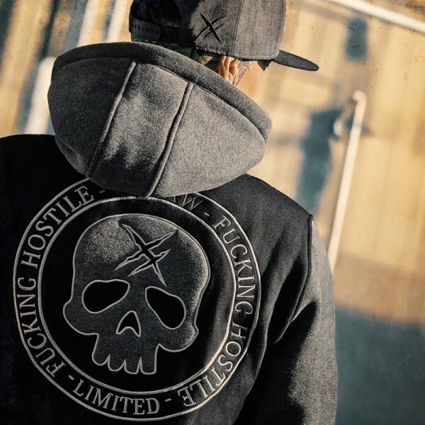 Photo en plan rapproché d'un homme de dos portant une casquette et un sweat zippé à capuche Hyraw Cloting. La photo met en valeur la broderie en double matière d’un énorme blason à tête de mort dans le dos de la veste. Le sweat est noir avec les manches, la capuche et les broderies grises qui rappellent les vestes type Varsity des universités américaines mais ici, dans un décor urbain, l'ambiance est résolument rock'n'roll à l’image de la marque de vêtements Hyraw Clothing.